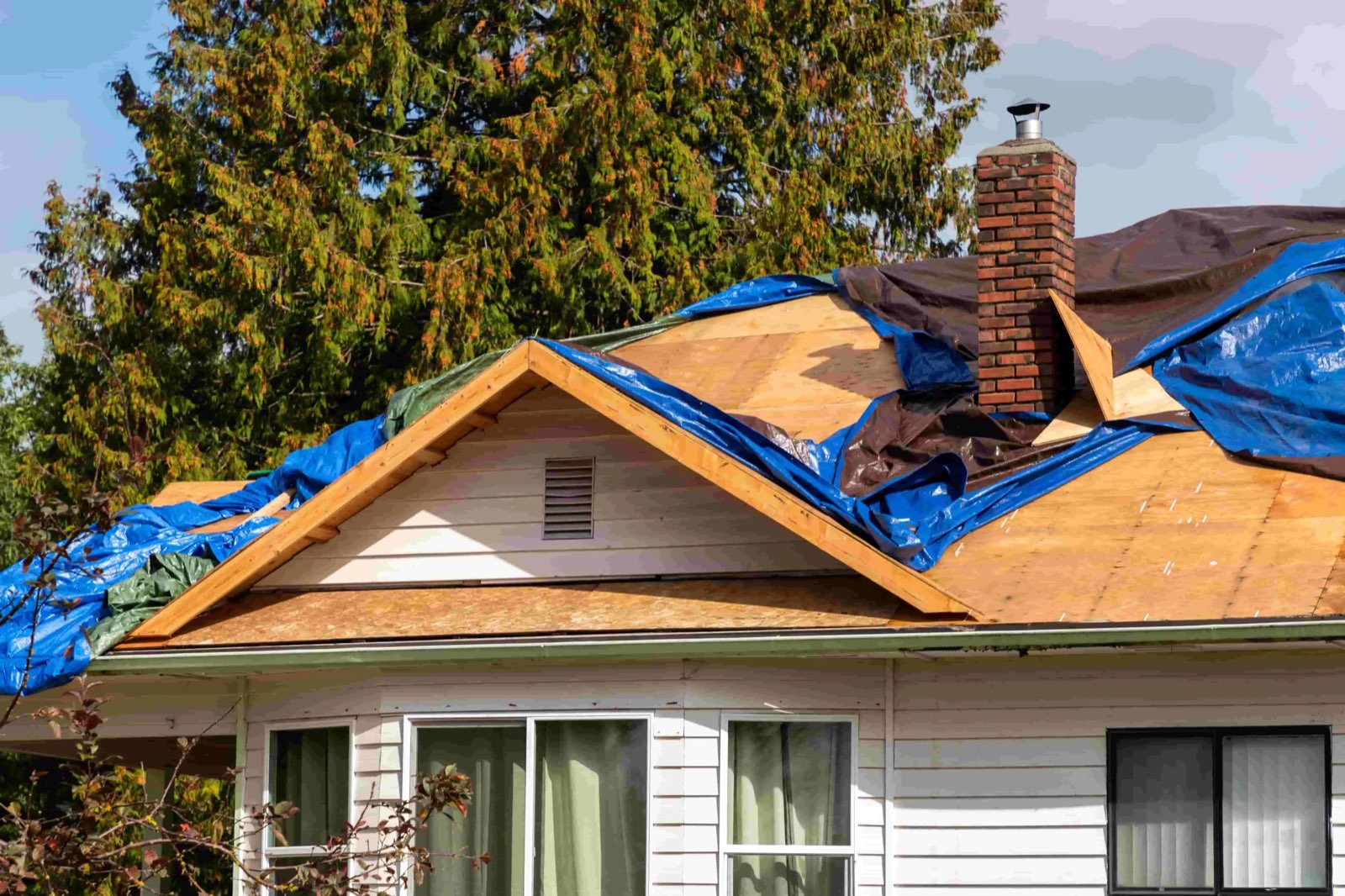 A storm-damaged house roof covered with blue and brown tarps over exposed sheathing around a brick chimney